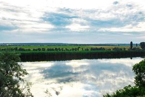 Summer evening dawn sky clouds reflected on a lake of water on the coast with green grass and forest trees photo