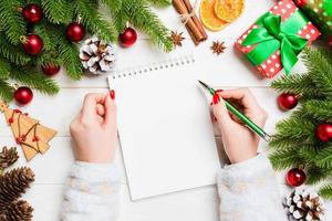 Top view of female hand writing in a notebook on wooden Christmas background. Fir tree and festive decorations. Wish list. New Year concept photo