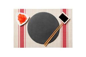 Emptyround black slate plate with chopsticks for sushi and soy sauce, ginger on sushi mat background. Top view with copy space for you design photo