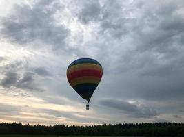 Large multi-colored bright round rainbow colored striped striped flying balloon with a basket against the sky in the evening photo