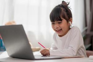 Asian schoolgirl doing her homework with laptop at home. Children use gadgets to study. Education and distance learning for kids. Homeschooling during quarantine. Stay at home photo