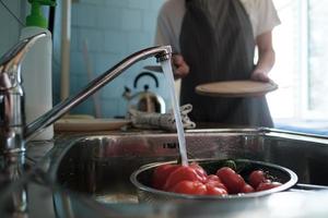 Fresh vegetables under running water on a colander in the sink, against the background of a wife with a knife and a cutting board. photo