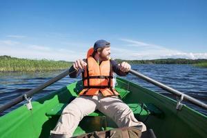 Contented bearded man in a life jacket rowing with oars on a boat and enjoying the beautiful scenery. Active lifestyle concept. photo