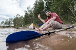 el hombre alegre descansa en la orilla del lago después de hacer kayak, hará un gesto victorioso y sonreirá mientras mira la cámara. foto