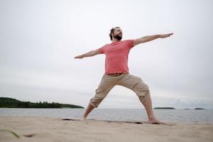 el hombre caucásico practica yoga, realiza ejercicios de virabhadrasana, de pie sobre una alfombra, en una playa de arena junto al lago. foto