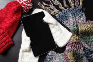 Tablet is on white mittens, as if in the hands of a person next to him, a knitted hat and a scarf are lying next to him. photo