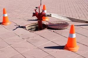 A large red metal fire hydrant with hoses and sleeves connected to the water source in the sewer hatch and emergency triangular orange cones warning of the danger photo
