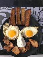 A merry smiling face, a face made of food, with eyes of fried eggs with yolks, a mouth of meat, nuggets, a nose of cheese curd, and hair from toast, bread on a black plate. Creative breakfast photo