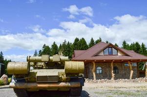 A large green military iron metal battle tank with a cannon is ridden parked next to the cottage house with a red tiled roof photo