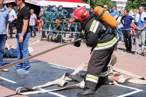 A fireman in a fireproof suit and a helmet running with an oxygen balloon pulling, holding a fire hose at a fire sport competition. Minsk, Belarus, 08.07.2018 photo