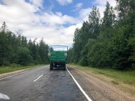 A truck, a tractor with a large green trailer is driving along a forest asphalt road with green trees on the grounds photo