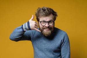 Funny shaggy-haired, bearded man wearing glasses and a sweater knitted making a hand gesture call me and looks camera. Yellow background. photo