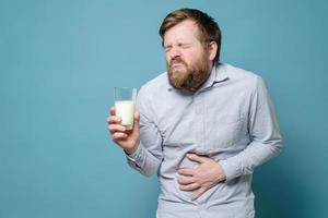 intolerancia a la lactosa. un hombre sostiene un vaso de leche en la mano y sufre un fuerte dolor abdominal. fondo azul. foto