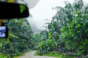 Rain drops on the car glass window with road in rainy season with forest, mountains and fog. photo