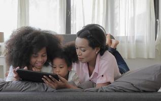 Modern family lifestyle concept. Young black american mother and her daughters are lay down on bed and looking to tablet device with enjoy feeling and happy smiling faces, copy space. photo