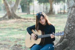 músicos asiáticos con una camisa negra de manga larga y gafas de sol en la cabeza. cantando y tocando la guitarra acústica debajo de un árbol en el parque por la mañana. foto