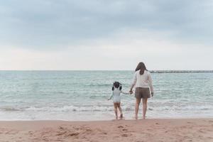 Asian cute girl and her mother walking or running or playing on the beach on summer holidays. Children with beautiful sea, sand and blue sky. Happy kids on vacations at seaside running on the beach. photo