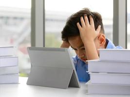 Young Asian elementary school boy sitting next to stack of books and using tablet computer with both hands holding his head with gestures and face showing stress. photo