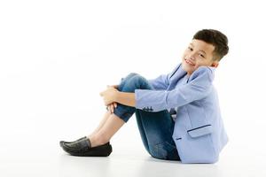 A 10-year-old Asian boy in a casual jacket is sitting on the ground, hands on his knees and smiling cheerfully, looking at the camera.