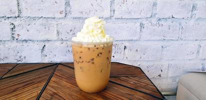 Plastic glass of iced coffee with whipped cream on top on brown wooden table with white rough wall background. Cold espresso, latte or cappuccino at caf. Drinking and food concept with copy space. photo