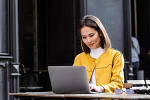 hermosa mujer asiática sentada con una computadora portátil net-book en un moderno bar de café, joven y encantadora trabajadora independiente asiática pensando en nuevas ideas durante el trabajo en una computadora portátil