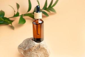 Natural medicine or aroma oil or beauty essence with dropper on stone podium stand with green ruskus branch on beige background. Face and body spa serum care concept photo