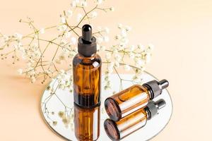 dos botellas de vidrio con cuentagotas para el cuidado de la piel facial en un espejo redondo con gypsophila blanca. cosmética natural. hidratante. fondo beige. foto