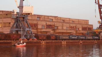 Sailboat moving at the Riga Freeport along the Daugava river, in Riga, Latvia. Port crane, container terminal and storage facility in the background. video