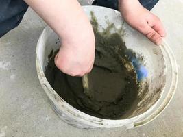 Builder hands with a metal spatula kneads in a large white plastic construction bucket plaster, tile glue, cement for the repair of an apartment, house, leveling the walls photo