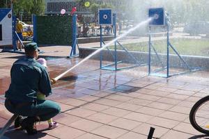 A fireman's man is teaching a child, a girl to put out a fire by shooting a stream of water from a bronzboyt, a fire hose, a hose to a target photo