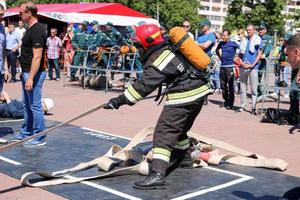 A fireman in a fireproof suit and a helmet running with an oxygen balloon pulling, holding a fire hose at a fire sport competition. Minsk, Belarus, 08.07.2018 photo