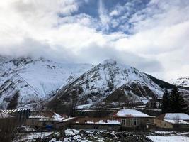 Small stone houses, buildings in the village on a beautiful mountain cold winter resort with high mountain peaks mist and snow covered rocks for snowboarding and skiing against a blue sky photo