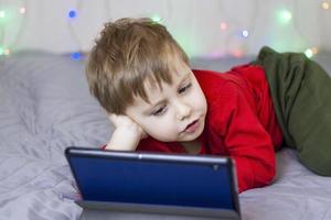 A cute boy is sitting on the bed and looking at a tablet. Funny smiling child. photo