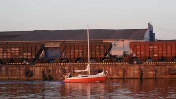 Sailboat moving at the Riga Freeport along the Daugava river, in Riga, Latvia. Port crane, container terminal and storage facility in the background. video