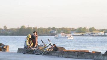 dos mujeres ciclistas hablando, sentadas, en el puerto franco de riga a lo largo del río daugava, en riga, letonia. transbordador pasando en segundo plano. video