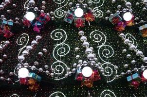 A fragment of a huge Christmas tree with many ornaments, gift boxes and luminous lamps. Photo of a decorated Christmas tree close-up