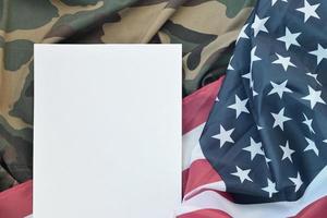 Blank paper lies on United States of America flag and folded military uniform jacket. Military symbols conceptual background banner and copy space photo