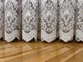 A curtain with a lace pattern barely touches the surface of the wooden floor. photo