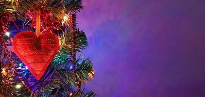 decoración hecha a mano de Navidad de corazón rojo con adorno en abeto en la noche de vacaciones, guirnalda de bokeh de colores, cuentas rojas, fondo púrpura oscuro borroso. banner de navidad con espacio de copia. foto