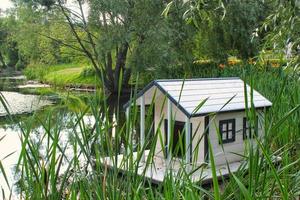 White wooden floating duck house in the centre of the pond in public park. Bird sanctuary in artificial lake. Spring nest. Summer green view. Wildlife nature reserve in river water. Waterfowl shelter photo