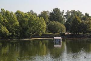 White wooden floating duck house in the centre of the pond in public park. Bird sanctuary in artificial lake. Spring nest. Summer green view. Wildlife nature reserve in river water. Waterfowl shelter photo