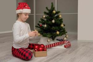 girl in pajamas and a santa hat sits next to the Christmas tree and chooses toys to decorate it photo