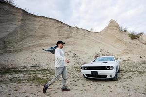 Handsome man in cap walking near his white muscle car in career. photo