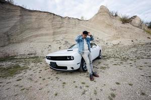 un hombre guapo con chaqueta de jeans y gorra está parado cerca de su auto blanco en carrera. foto