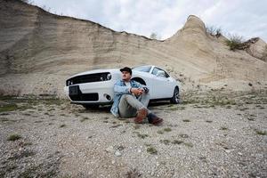 un hombre guapo con chaqueta de jeans y gorra está sentado cerca de su auto blanco en carrera. foto