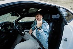 un hombre guapo con chaqueta de jeans y gorra se sienta en su auto blanco y muestra el signo de la mano del rock and roll. foto