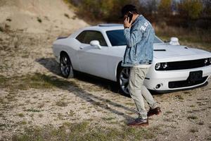 hombre guapo con chaqueta de jeans y gorra llamando por teléfono mientras está parado cerca de su auto blanco en carrera. foto