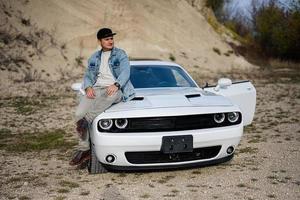 un hombre guapo con chaqueta de jeans y gorra se sienta en el capó de su auto blanco en carrera. foto