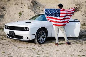 parte trasera del hombre con la bandera de estados unidos cerca de su muscle car americano blanco en carrera. foto