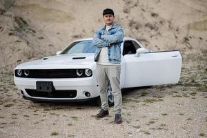 Handsome man in jeans jacket and cap is standing near his white muscle car in career. photo